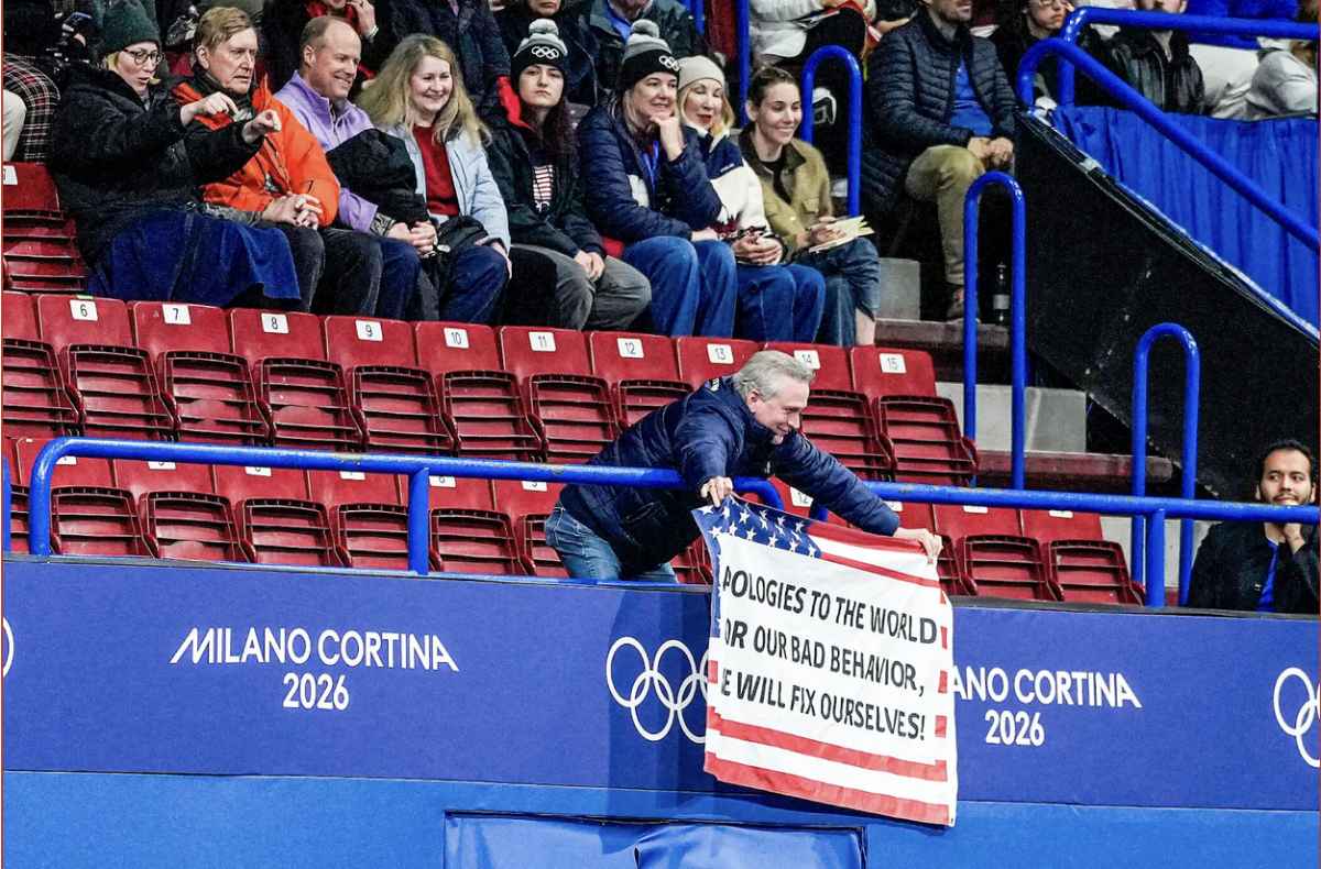 A spectator at an Olympic figure skating event held a banner of the United States flag with a message of apology. Image Credit NYT.